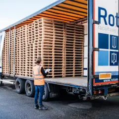 Pracownik sprawdza, ile pustych palet wchodzi na naczepę. Stosy drewnianych palet czekają na załadunek.