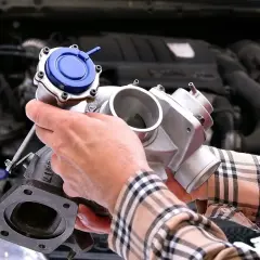 Ręce mechanika trzymające turbosprężarkę z niebieskim zaworem przed silnikiem samochodu.