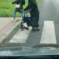 Starsza pani z chodzikiem i młodsza osoba przechodzą przez przejście dla pieszych.