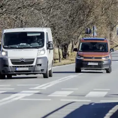 Biała furgonetka wyprzedza na przejściu dla pieszych pomarańczowego vana. W tle stacja benzynowa.