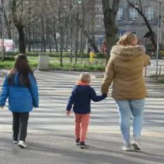 Rodzina przechodzi przez pasy. Kobieta trzyma za rękę małego chłopca, a obok idzie starsza dziewczynka. Szczególna ostrożność kiedy przechodzimy przez jezdnię.
