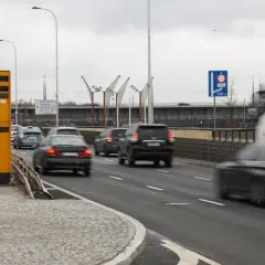 Żółty fotoradar stoi przy drodze, gdzie ruch jest duży. Znak informuje, od ilu łapie fotoradar, a samochody pędzą obok.