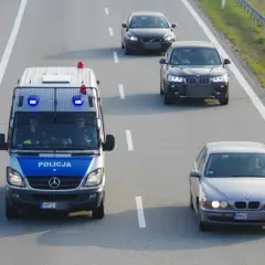 Policyjny van z włączonymi sygnałami świetlnymi jedzie autostradą, wyznaczając ile minimum na autostradzie. Obok jadą inne samochody.