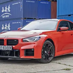 Pomarańczowe BMW M2 z czarnymi felgami, stojące przed niebieskimi kontenerami. Sprawdź aktualną bmw m2 cena!