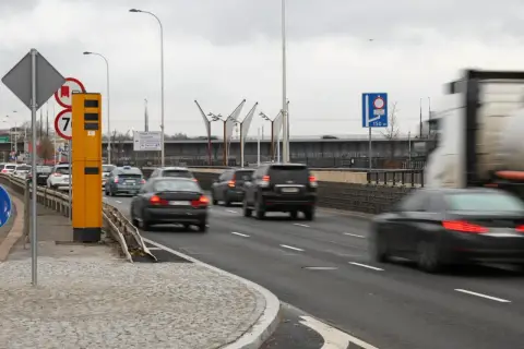 Żółty fotoradar stoi przy drodze, gdzie ruch jest duży. Znak informuje, od ilu łapie fotoradar, a samochody pędzą obok.