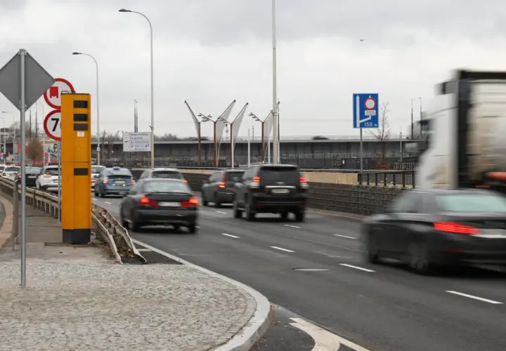 Żółty fotoradar stoi przy drodze, gdzie ruch jest duży. Znak informuje, od ilu łapie fotoradar, a samochody pędzą obok.