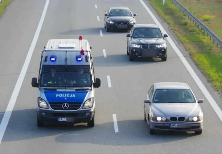 Policyjny van z włączonymi sygnałami świetlnymi jedzie autostradą, wyznaczając ile minimum na autostradzie. Obok jadą inne samochody.