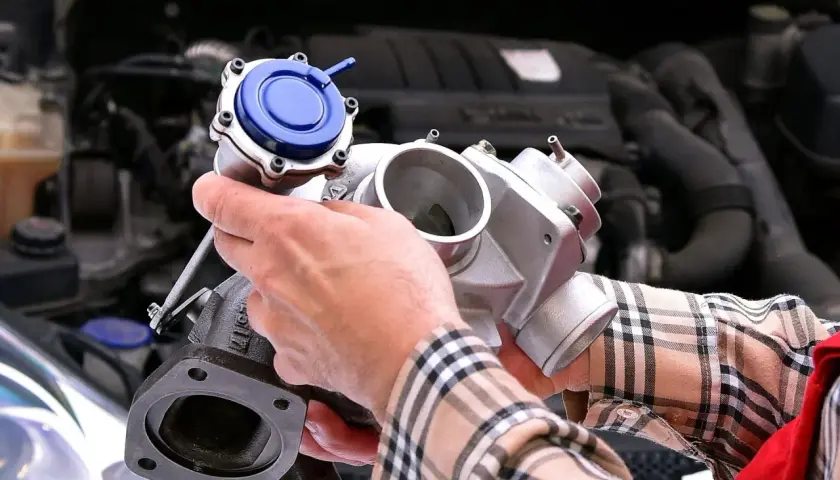Ręce mechanika trzymające turbosprężarkę z niebieskim zaworem przed silnikiem samochodu.