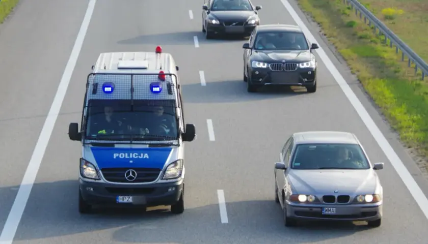 Policyjny van z włączonymi sygnałami świetlnymi jedzie autostradą, wyznaczając ile minimum na autostradzie. Obok jadą inne samochody.