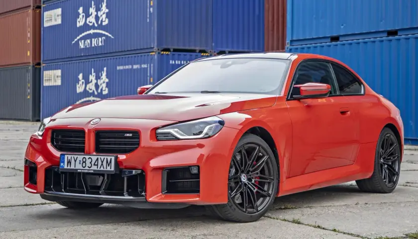 Pomarańczowe BMW M2 z czarnymi felgami, stojące przed niebieskimi kontenerami. Sprawdź aktualną bmw m2 cena!