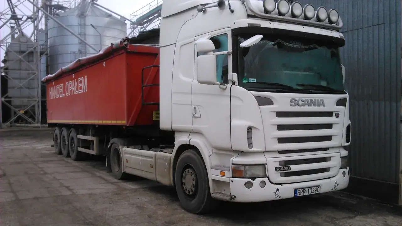 Biała Scania R420 z naczepą "Handel Opałem" stoi przy silosach. Zastanawiasz się, ile kosztuje nowa Scania?