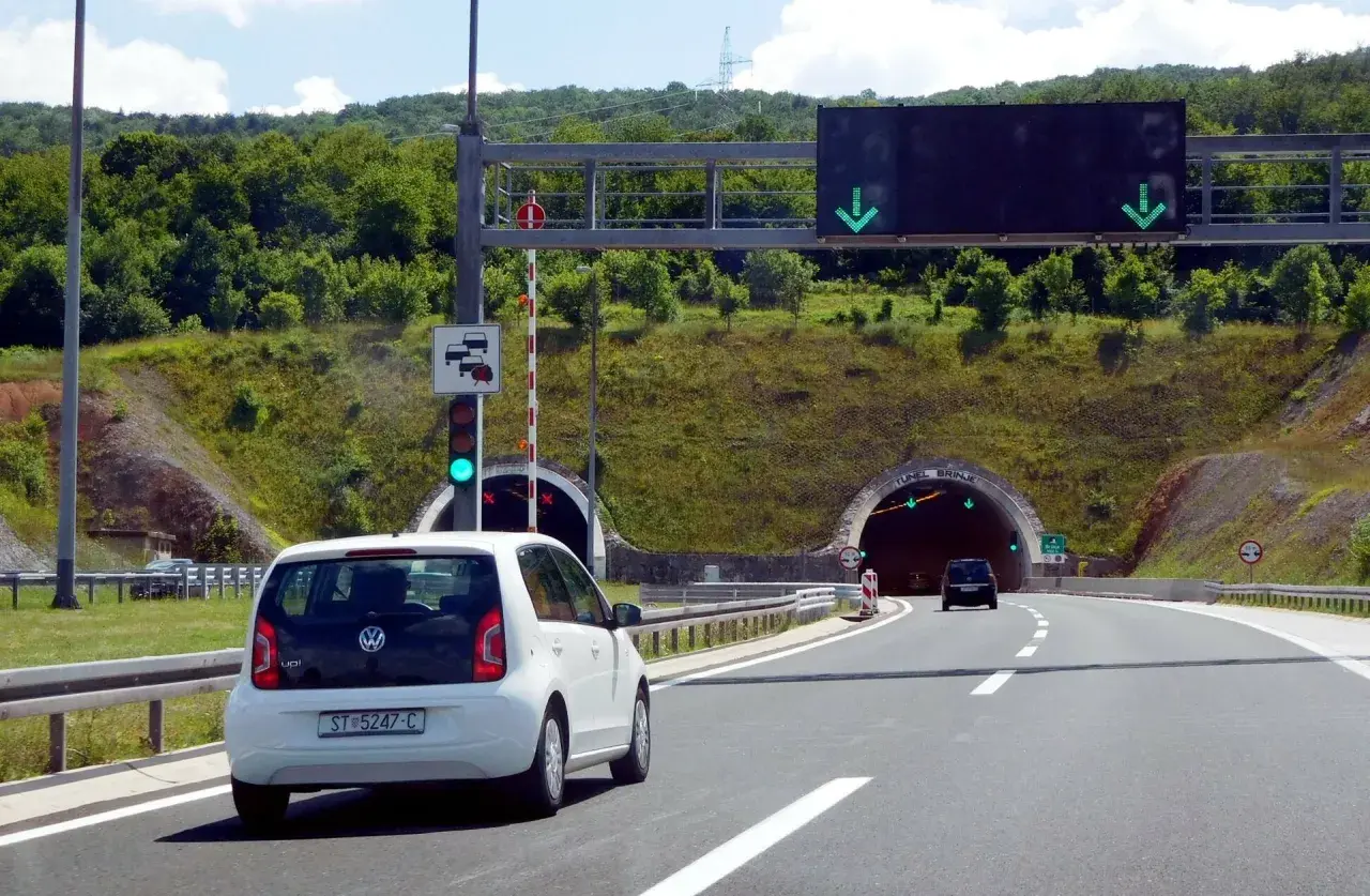 Biały samochód zbliża się do tunelu w Chorwacji. Znak drogowy informuje o opłatach za autostrady.