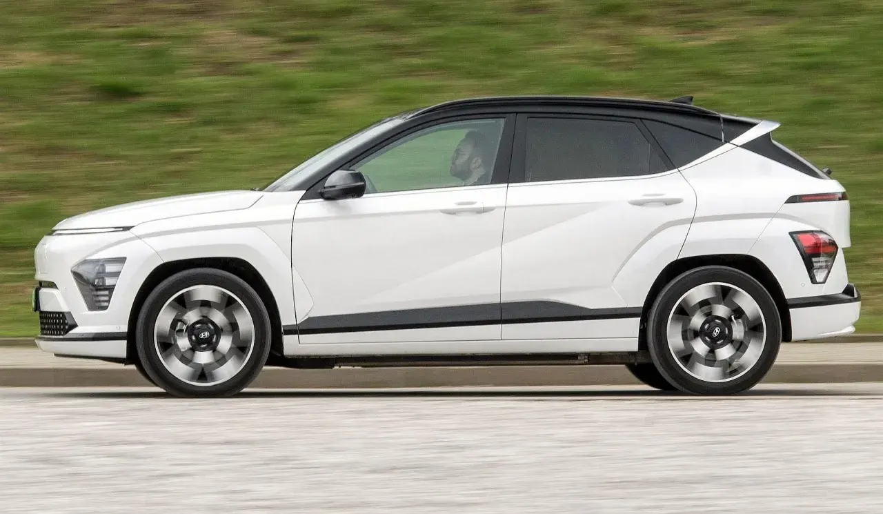 Biały Hyundai Kona w ruchu, z czarnym dachem i przyciemnianymi szybami. Kierowca widoczny w kabinie. Sprawdź hyundai kona wymiary.