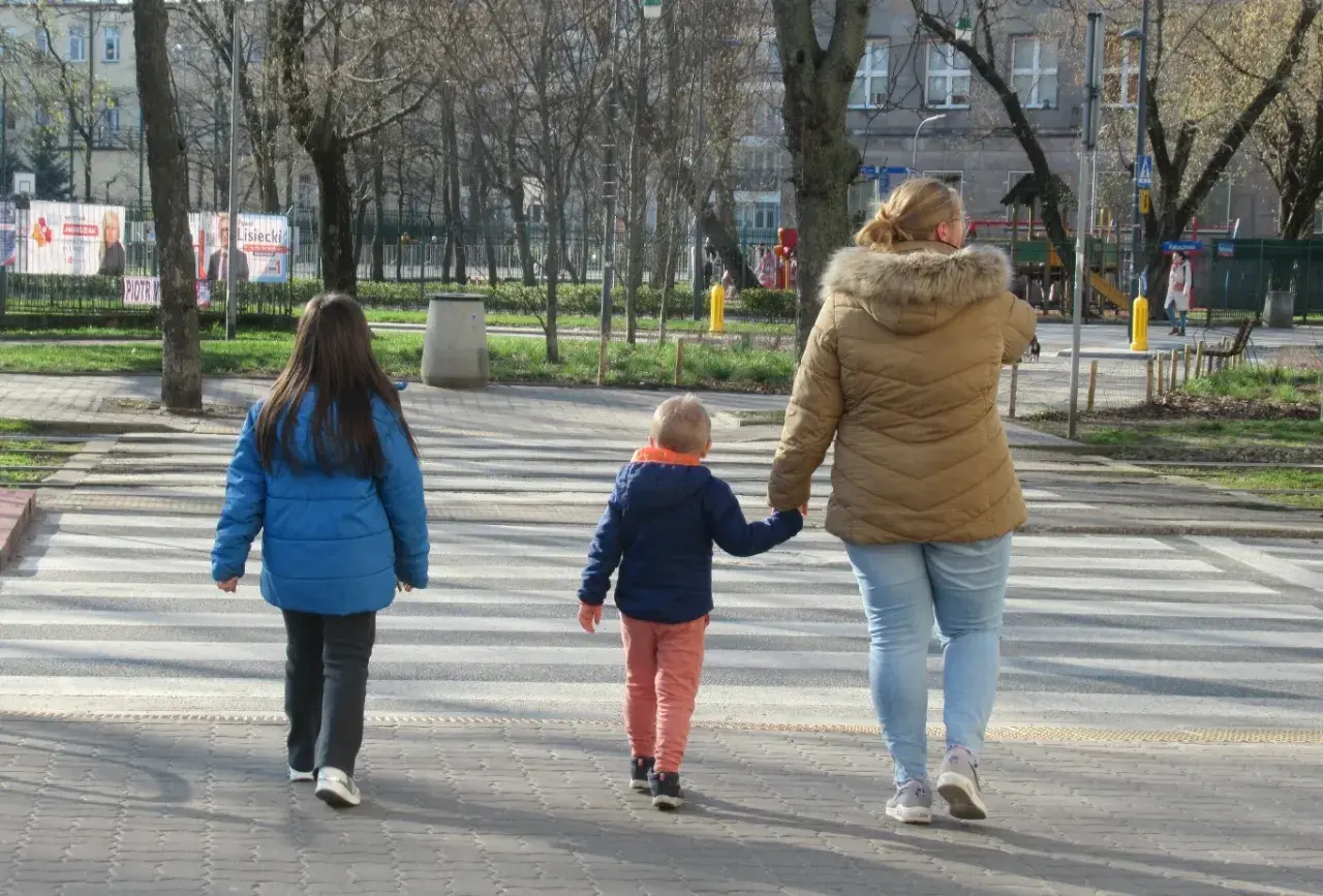 Rodzina przechodzi przez pasy. Kobieta trzyma za rękę małego chłopca, a obok idzie starsza dziewczynka. Szczególna ostrożność kiedy przechodzimy przez jezdnię.