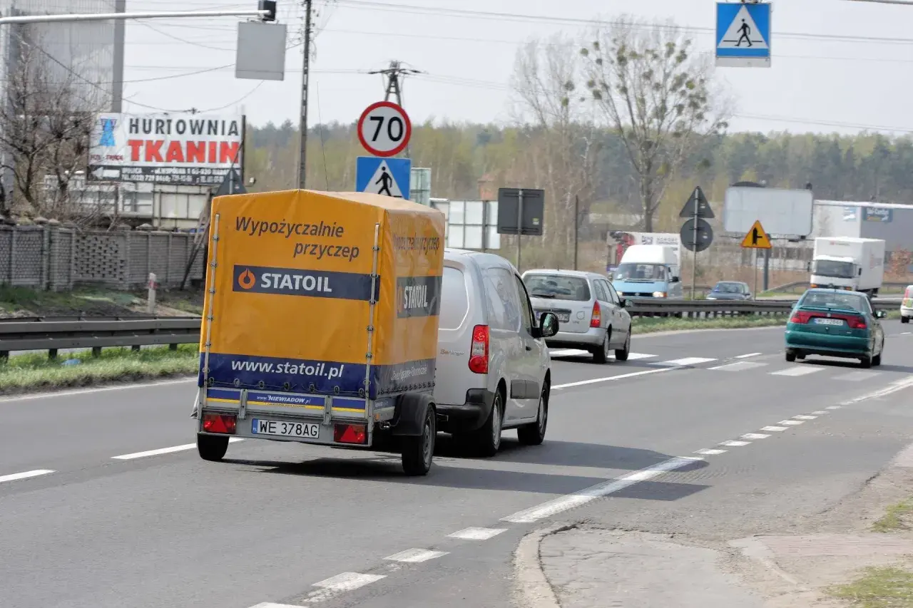 Na drodze widać przyczepę z napisem "Wypożyczalnie przyczep Statoil". Znak ograniczenia prędkości wskazuje 70 km/h, co sugeruje, ile autobus może jechać na autostradzie w takich warunkach.