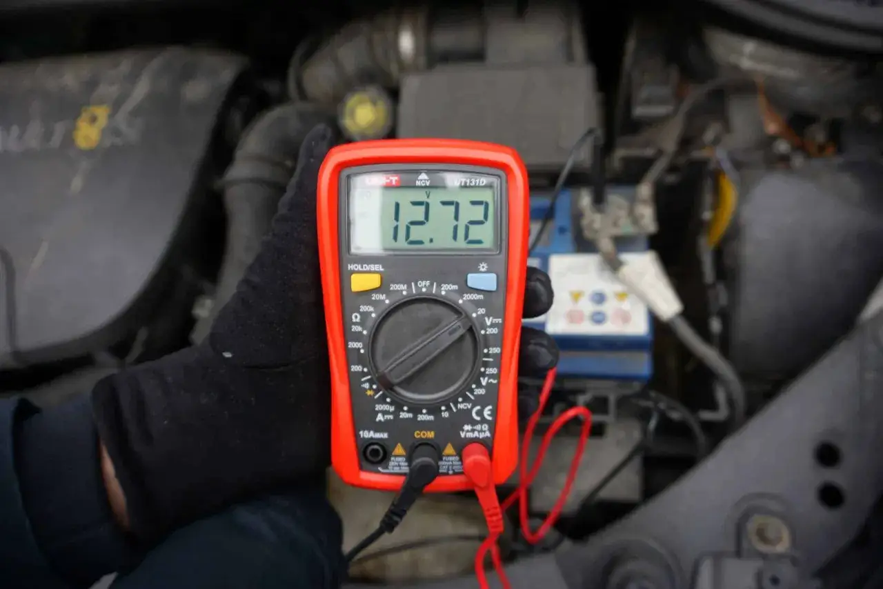 Ręka w czarnej rękawiczce trzyma multimetr pokazujący 12.72V, co oznacza, że akumulator jest w pełni naładowany.