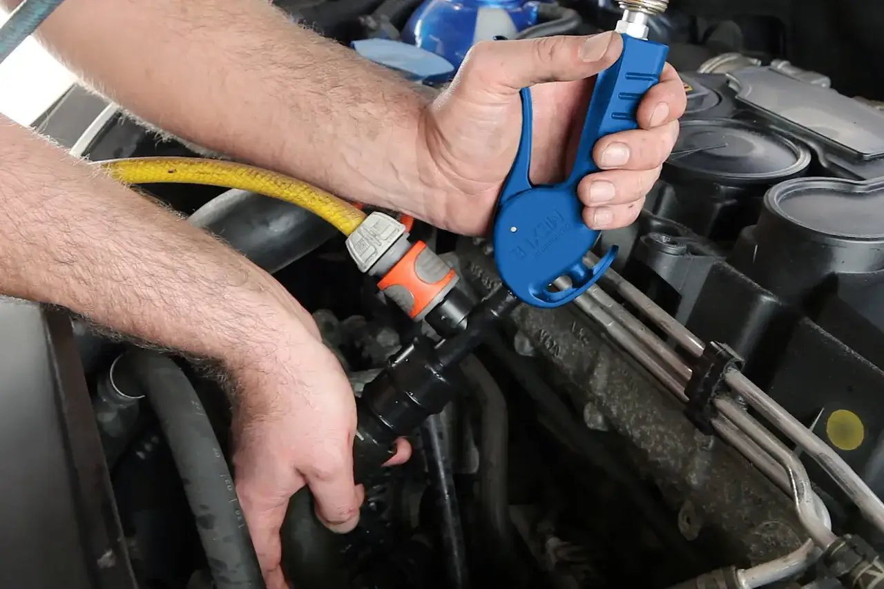 Ręce mechanika podłączają niebieskie narzędzie do węża, by odkamienić układ chłodzenia w samochodzie.