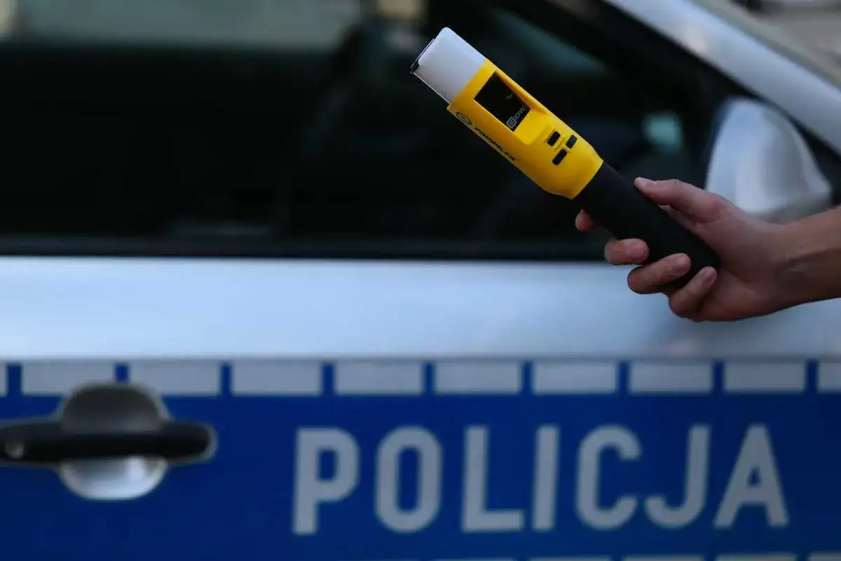 Dłoń trzyma żółty alkomat na tle radiowozu policyjnego. Czy na komendzie można zbadać się alkomatem? Tak, policja używa takich urządzeń.